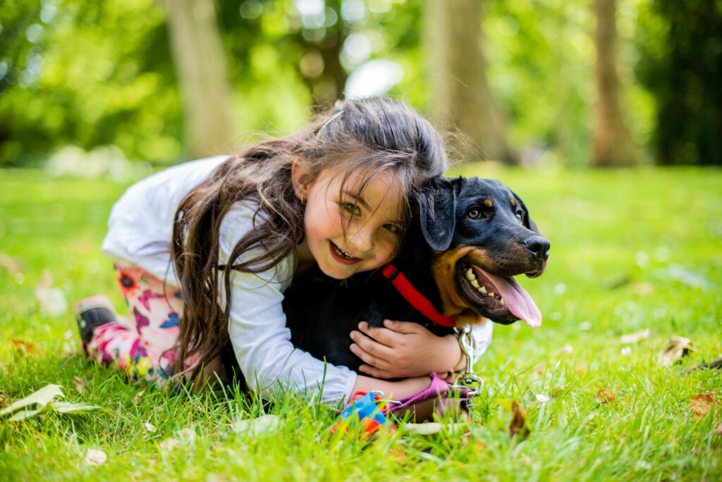 dziewczyna w różowej kurtce bawi się czarnym i brązowym psem o krótkiej powłoce na zielonym polu trawy